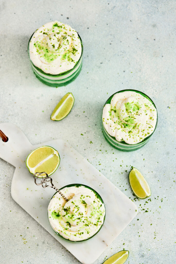 Three glasses of creamy Jello Parfaits, topped with lime zest, sit on a light surface with lime wedges and a marble board holding a slice of lime and a small whisk.
