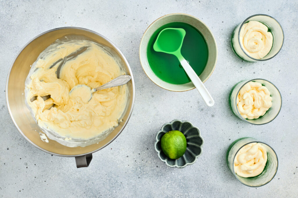 A mixing bowl with creamy batter, a bowl of green liquid for St. Patrick’s Day Green Jello Parfaits, a lime, and three glass cups filled with swirled batter are arranged on a light surface.