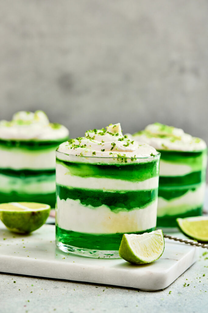A festive St. Patrick’s Day Green Jello Parfait featuring layers of green jelly and white cream, topped with whipped cream, green sprinkles, and garnished with lime wedges—a perfect treat for your celebration!.