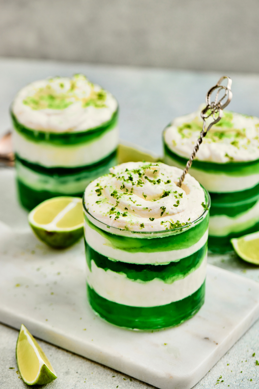 Three glass cups containing layered green jelly and white cream, topped with lime zest, are placed on a marble board with lime wedges nearby—perfect as festive St. Patrick’s Day Green Jello Parfaits.