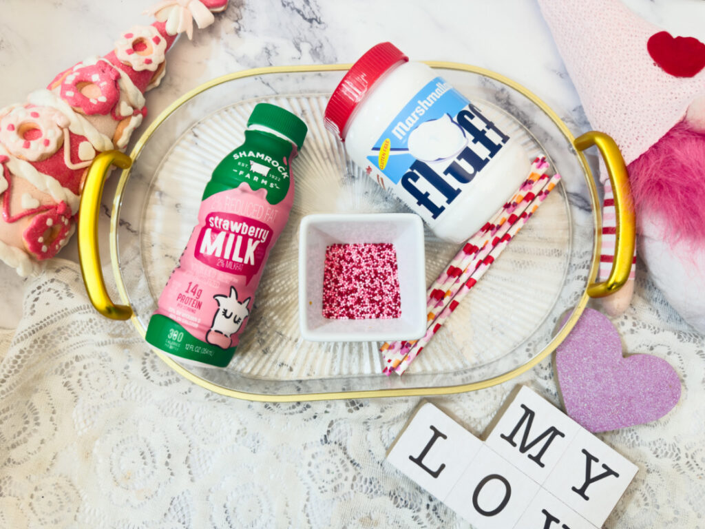 A glass tray holds Valentine’s Fluff Drink made with strawberry milk and marshmallow fluff, pink sprinkles in a dish, two striped straws, and sits among heart and love-themed decorations.