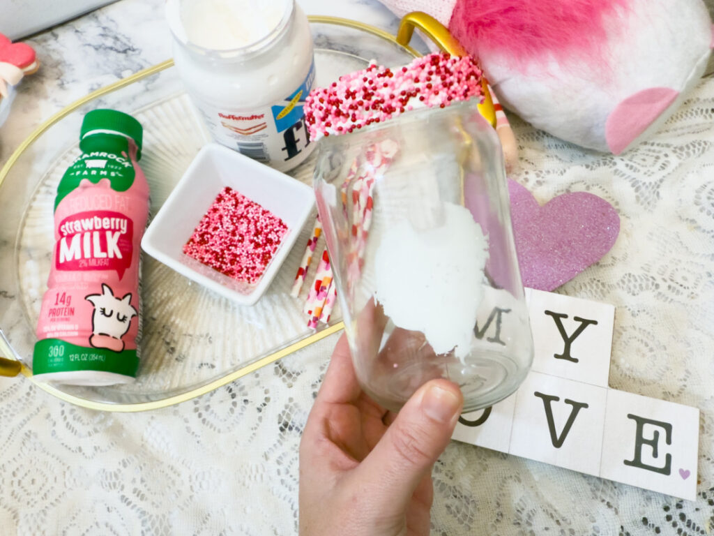 A hand holds a glass jar decorated with a white heart and red sprinkles, ready for Strawberry Milk, near bottles, a bowl of sprinkles, and blocks spelling "MY LOVE" on a lace tablecloth.