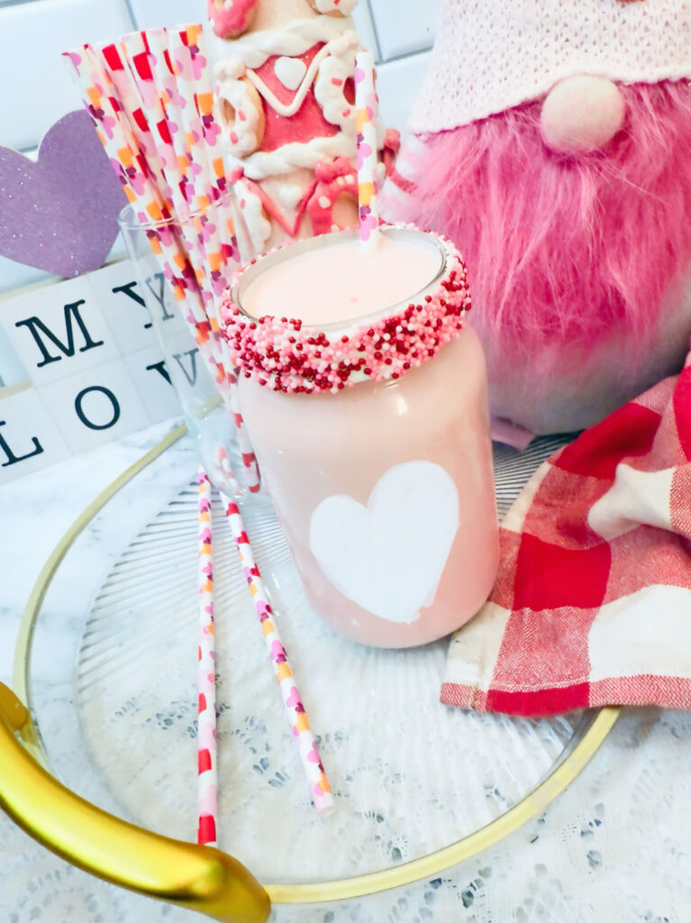 A mason jar filled with strawberry milk, topped with red and white sprinkles, surrounded by colorful straws and Valentine-themed decorations—a perfect Valentine’s treat.