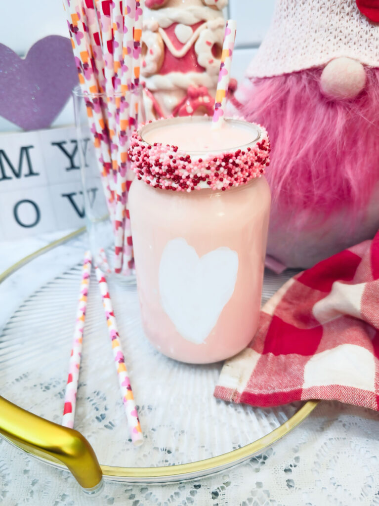 A glass jar of Valentine’s Fluff Drink—pink strawberry milk with a white heart, pink sprinkles, and straw—sits on a tray with decorative straws and a pink gingham napkin, making it the perfect Valentine treat.