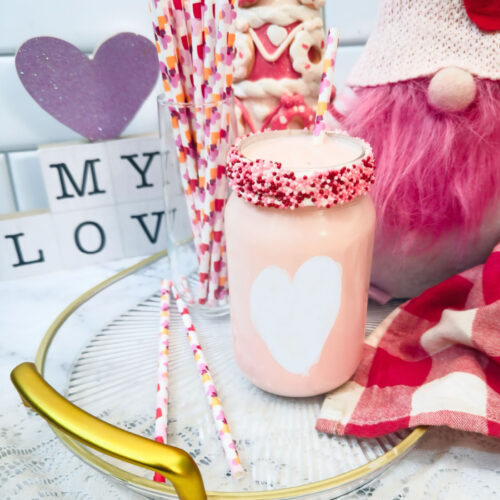 A pink Valentine’s Fluff Drink in a jar with a heart design, surrounded by decorative straws, a gnome doll, and Valentine’s-themed ornaments on a tray—an irresistible strawberry milk treat for the season of love.