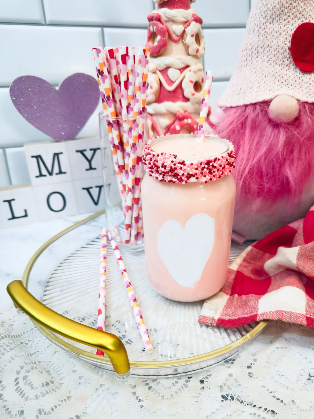 A pink Valentine’s Fluff Drink in a jar with a heart design, surrounded by decorative straws, a gnome doll, and Valentine’s-themed ornaments on a tray—an irresistible strawberry milk treat for the season of love.