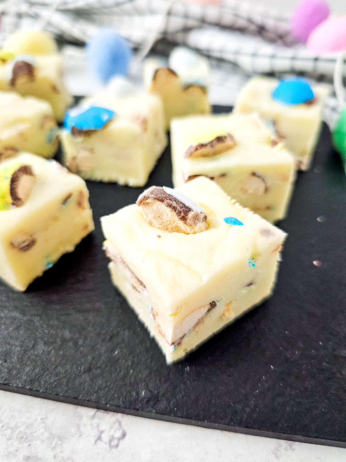 Squares of Robin Eggs Fudge, studded with candy and nut pieces, are artfully arranged on a black slate surface.